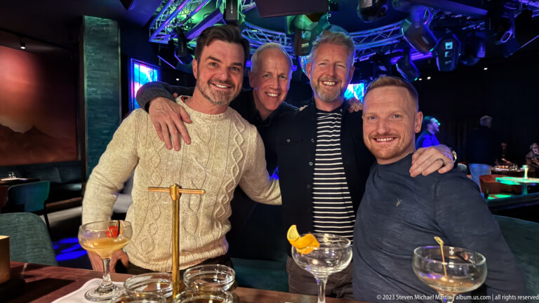 Four men pose behind a table with cocktails and glassware under blue and green lights in a lively bar or club setting.