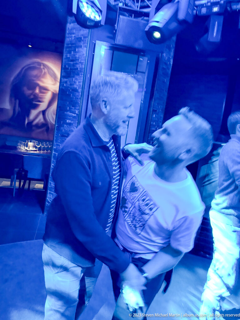 Two men face each other closely on a blue-lit nightclub dance floor, surrounded by other patrons.