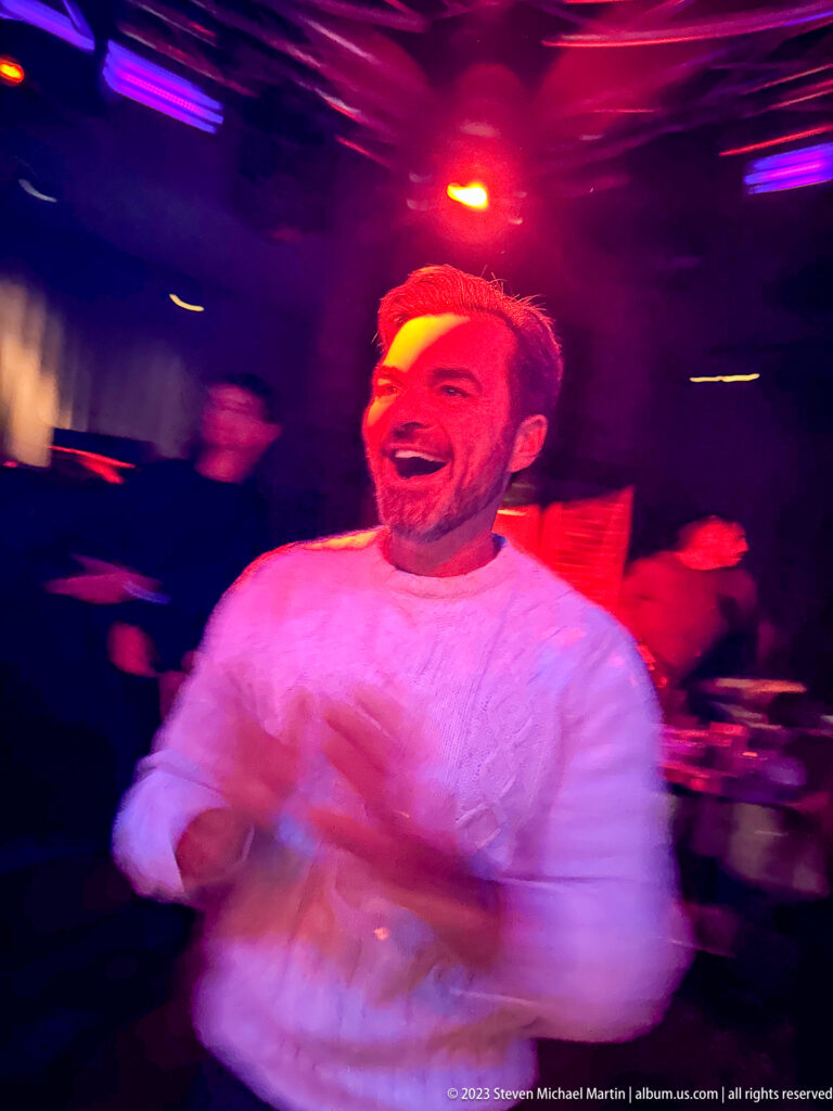 Laughing man in a light cable-knit sweater moving under red and magenta nightclub lighting.