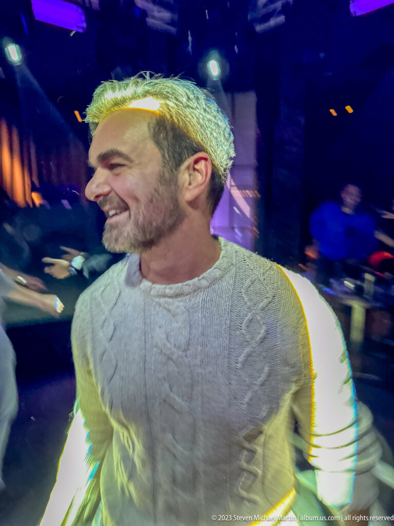 Smiling man in a pale cable-knit sweater under mixed yellow, green, and violet nightclub lighting.