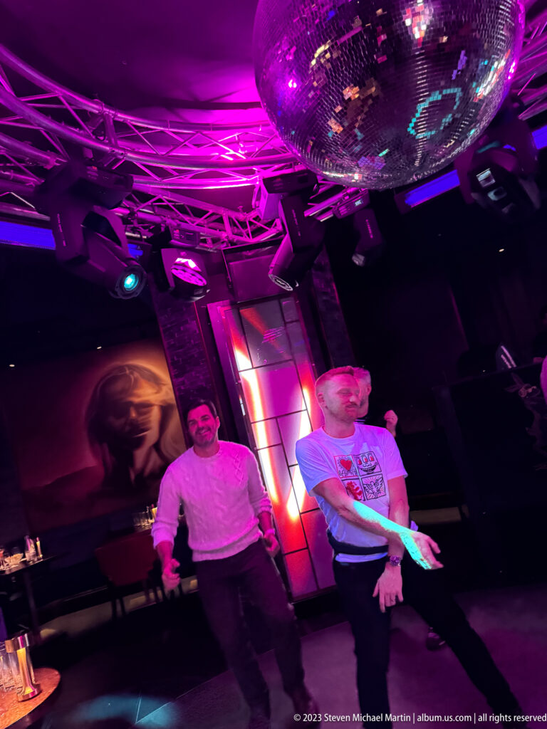 Two men stand beneath a large mirrored disco ball and lighting truss in a purple-lit nightclub.