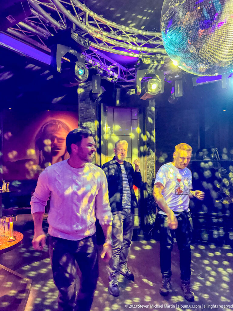 Three men on a nightclub dance floor beneath a mirrored disco ball, with yellow reflected light spots across the room.