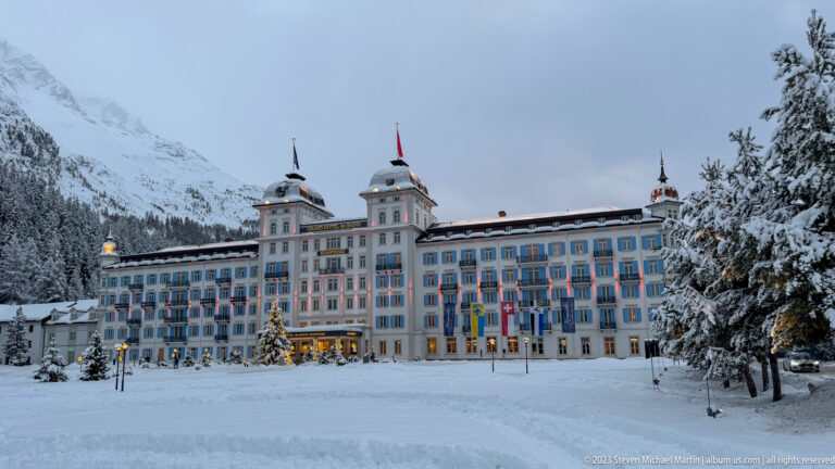 Grand Hotel des Bains Kempinski stands behind deep snow, with flags, blue shutters, towers, and a lit Christmas tree.