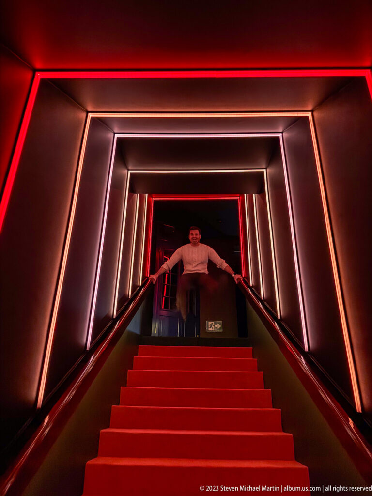 Red-carpeted staircase framed by glowing rectangular light strips, with a man standing at the top landing.