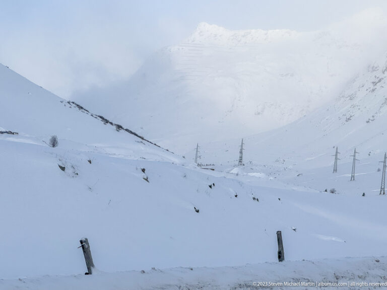 Snow-covered alpine landscape with a road, utility poles, and mountains fading into low cloud and blowing snow.