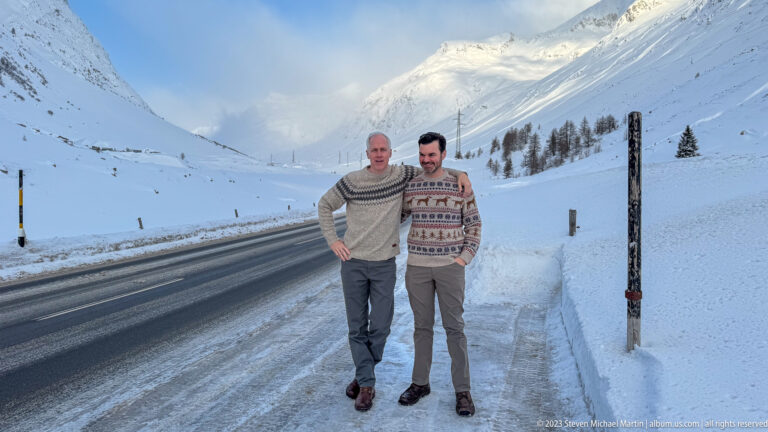 Two men in patterned sweaters pose beside a snow-lined alpine road with mountains rising behind them.