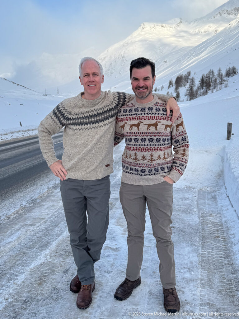 Two men in patterned knit sweaters pose together beside a snowy alpine road and mountain slope.