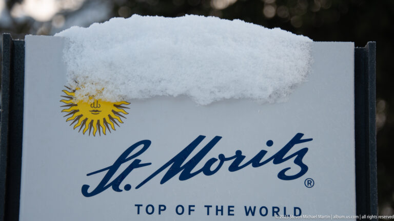 Snow-covered sign reading 'St. Moritz Top of the World' with the yellow sun emblem and blue script logo.