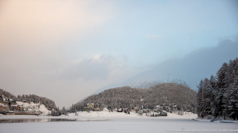Frozen Lake St. Moritz with snowy wooded hills, scattered buildings, and low pastel cloud over the distant mountains.