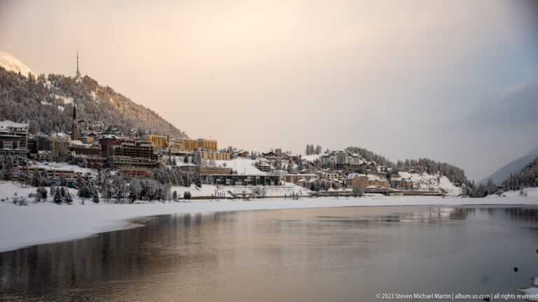 Snowy St. Moritz hillside above dark open lake water reflecting buildings beneath a pale peach winter sky.