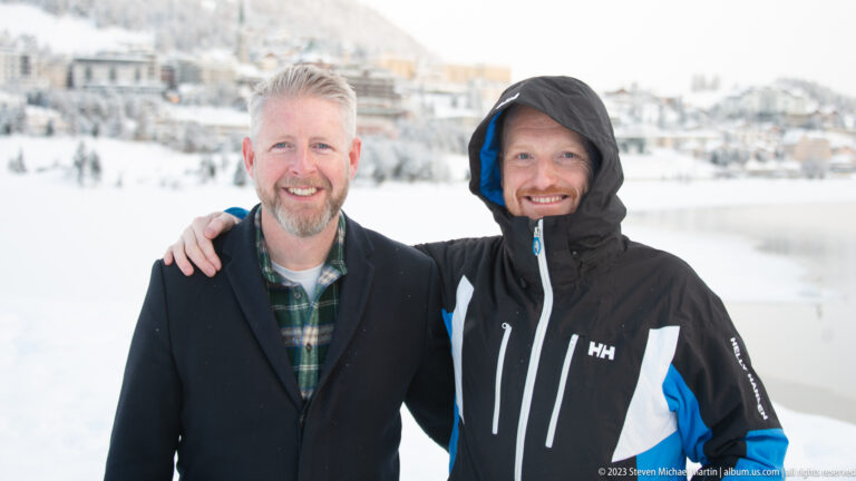 Two men pose together beside snowy Lake St. Moritz, with resort buildings softly blurred in the background.