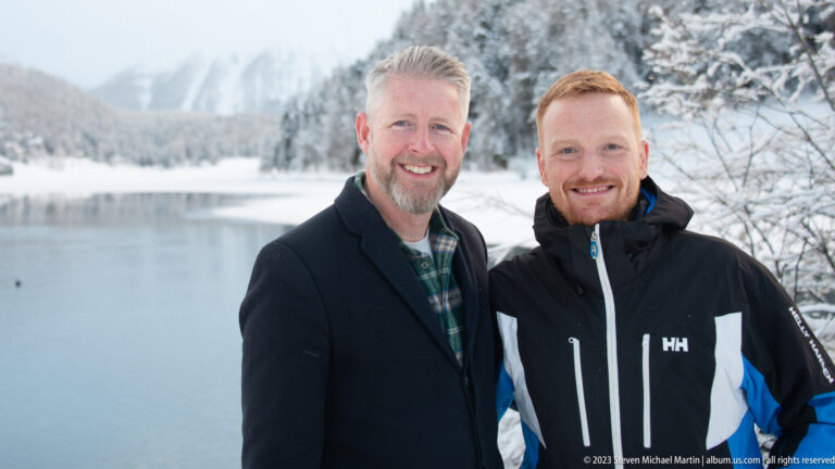 Two men pose beside calm winter lake water with snowy trees and shoreline behind them.