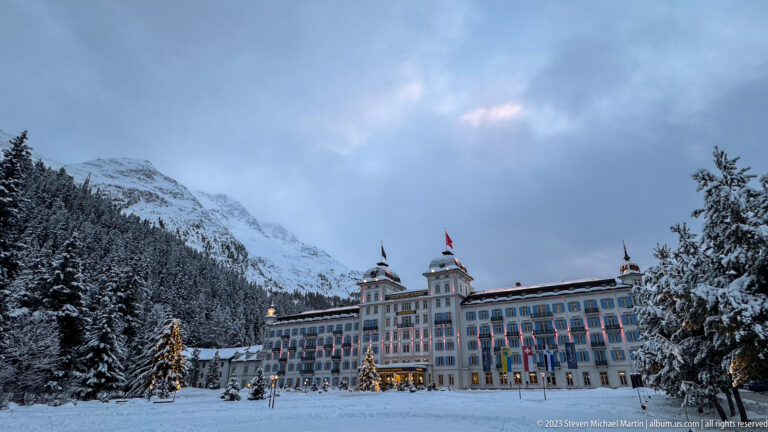 Grand Hotel des Bains Kempinski sits beneath a snowy mountain, with a lit Christmas tree in front.