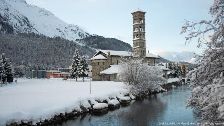 Snow-covered stone church with a tall multi-stage bell tower beside water and wooded mountains.