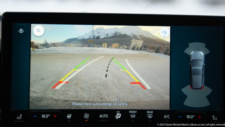 Vehicle dashboard display showing a rear-view camera with parking guidelines, sensor graphic, snow, and mountains behind.