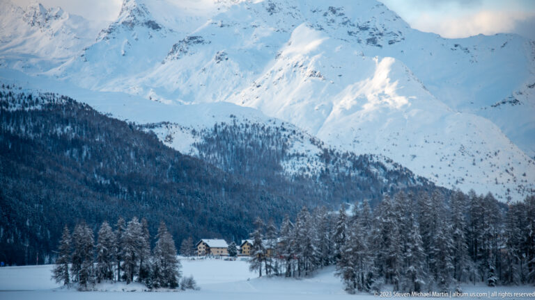 Snow-covered mountain massif above dark conifer forest and a small cluster of alpine houses.