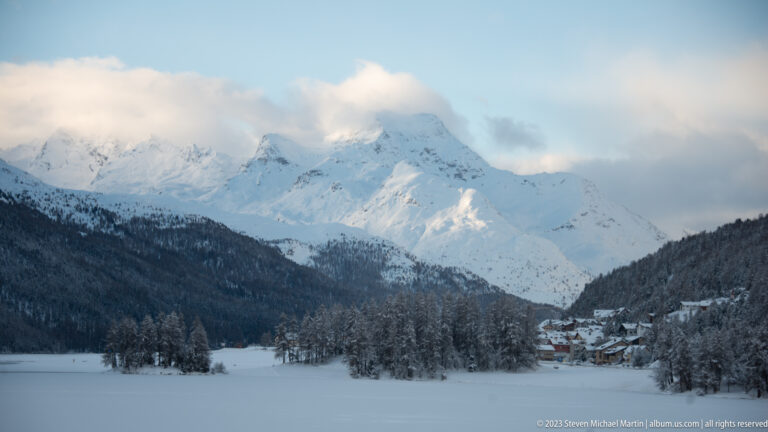 Snow-covered alpine peaks above a forested valley and small settlement, with clouds drifting around the summits.