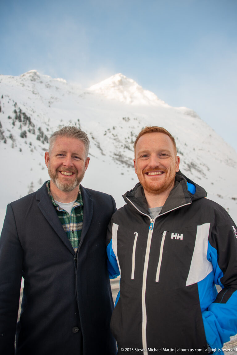 Two smiling men pose together in front of a broad snowy mountain slope under a clear blue sky.