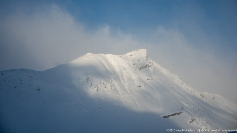 Sunlit snow-covered mountain ridge emerging from deep blue shadow, with cloud or windblown snow along the crest.