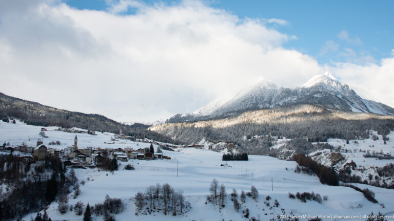Snow-covered alpine village and open valley beneath a forested mountain, with sunlight breaking through clouds.