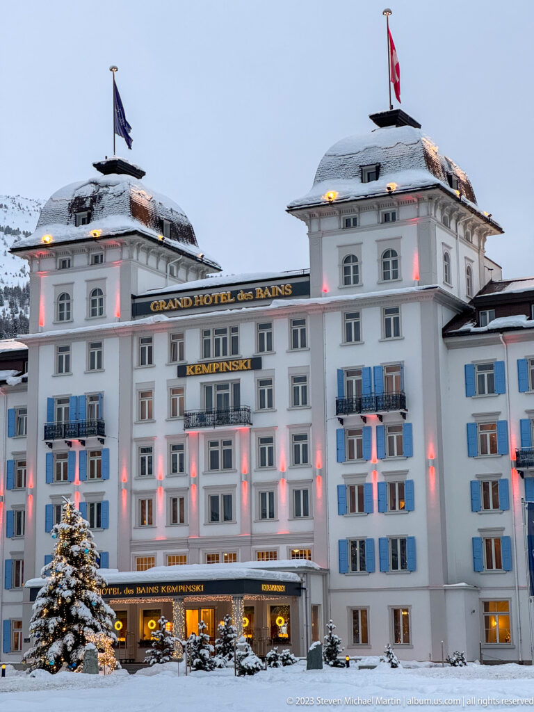 Central façade of the Grand Hotel des Bains Kempinski with visible signage, blue shutters, snow, and red winter lighting.