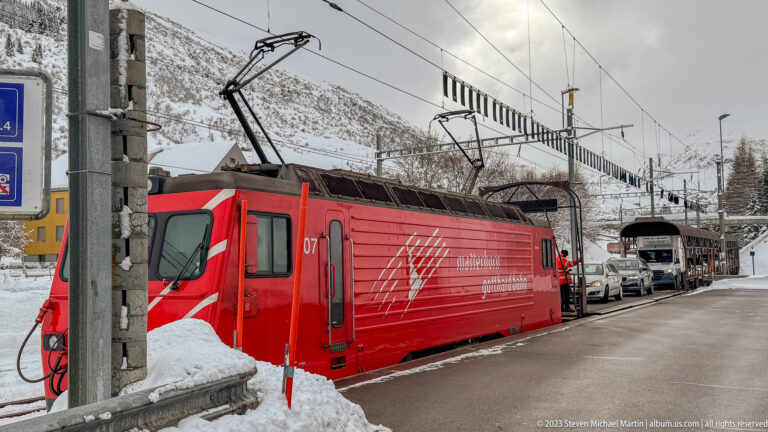 Red Matterhorn Gotthard Bahn electric locomotive under overhead wires beside snowbanks, with vehicles and another rail vehicle behind.
