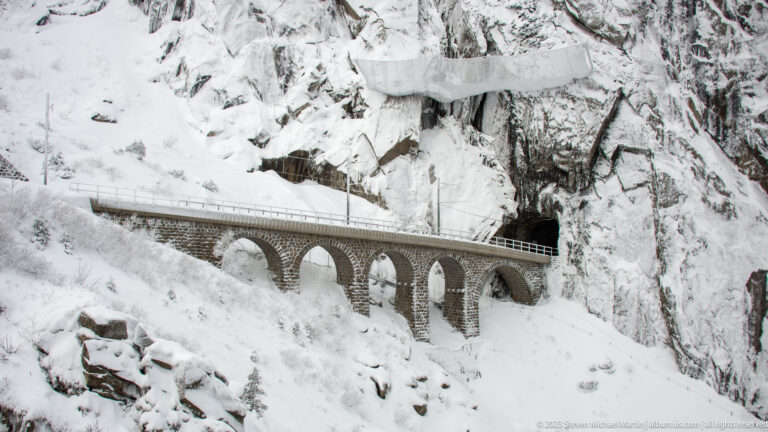 Stone viaduct with multiple rounded arches crossing a snowy ravine and entering a tunnel in a rock cliff.