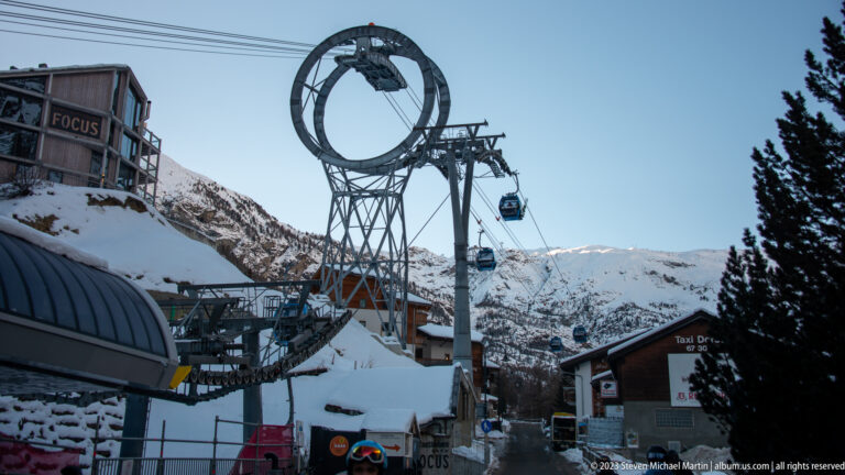 Gondola cabins passing a snowy cableway station with a large circular support structure and mountain slopes behind.