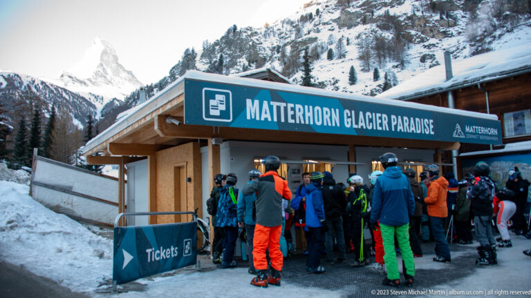 Crowd of skiers and visitors beneath a large MATTERHORN GLACIER PARADISE sign at a snowy ticket building.