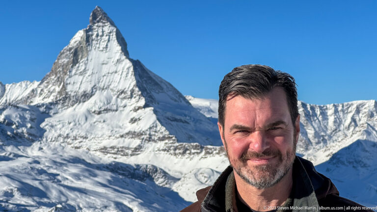 Dark-haired man in a brown winter jacket posing with the Matterhorn behind him under a clear blue sky.