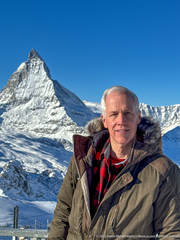 Gray-haired man in a fur-trimmed parka and red plaid scarf posing with the Matterhorn.