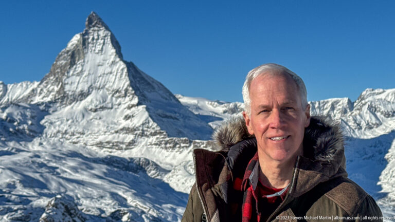 Close portrait of a gray-haired man in a fur-trimmed parka with the Matterhorn behind him.