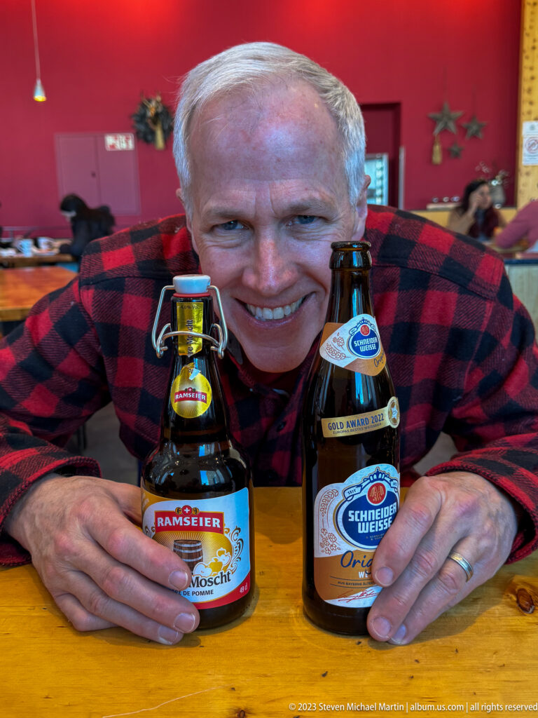 Smiling gray-haired man leaning between two swing-top Swiss beer bottles on a wooden table.