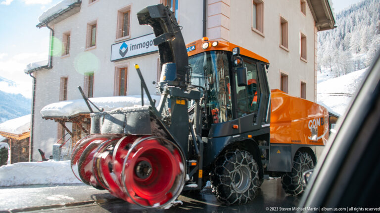 Orange snow-removal vehicle with large red rotary blower and traction-equipped tires beside deep roadside snow.