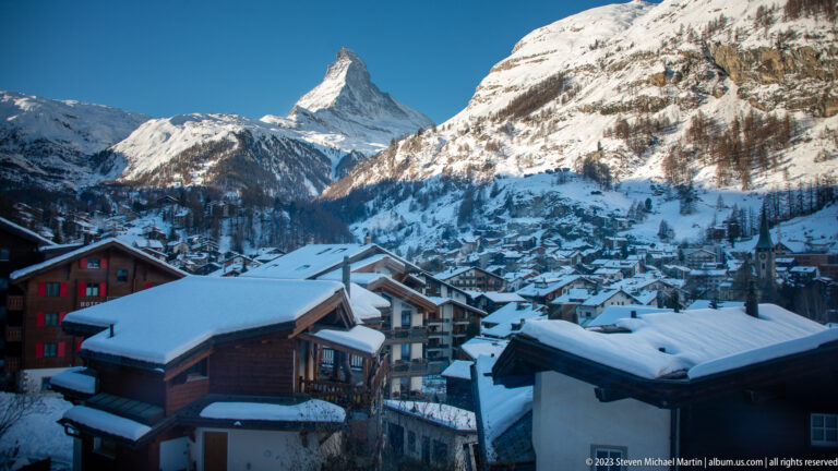Snow-covered Zermatt rooftops and chalets below the distant Matterhorn and steep mountain slopes.