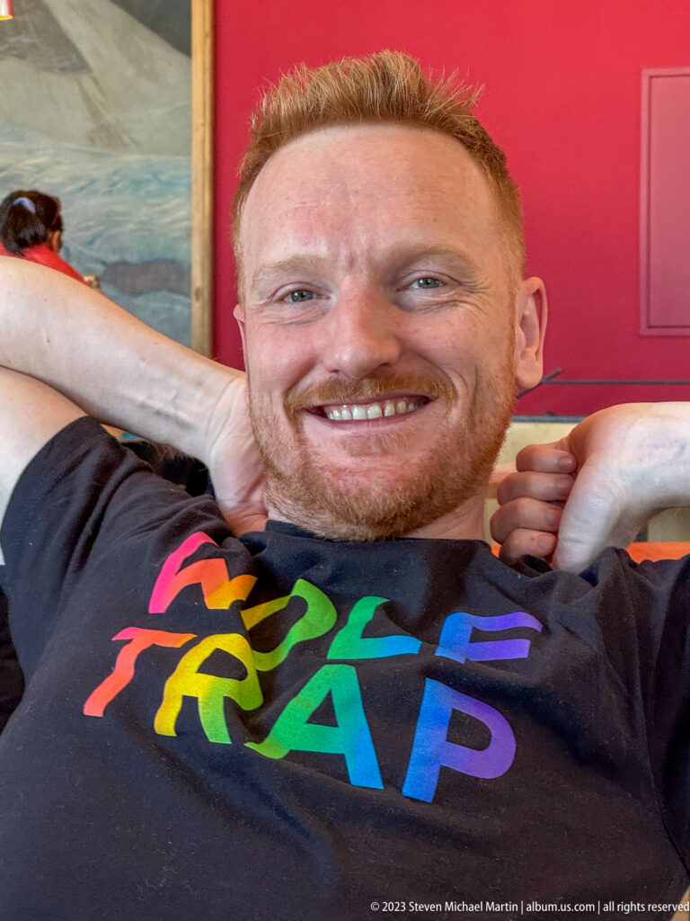 Smiling red-haired man reclining indoors in a black T-shirt with large rainbow-colored lettering.
