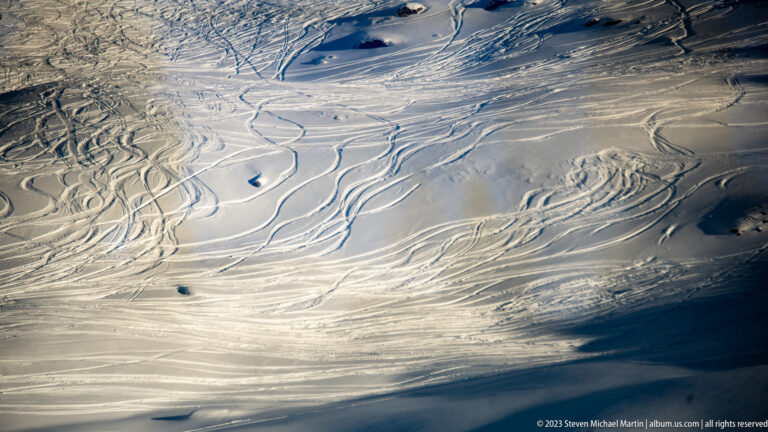 Broad snowfield densely crossed by overlapping ski and snowboard tracks and long blue shadows.