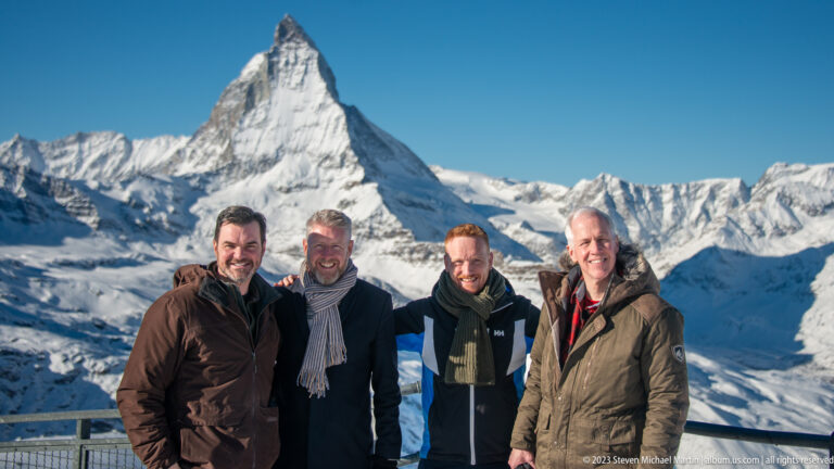 Four men in winter coats posing together with the snow-covered Matterhorn directly behind them.