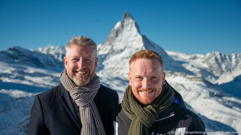 Two smiling men in scarves and winter coats posing with the Matterhorn softly focused behind.
