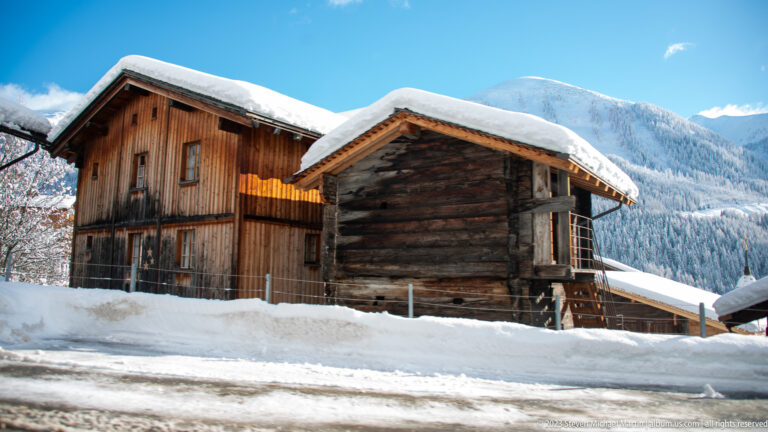 Two traditional timber buildings with snow-covered pitched roofs beside a road, backed by a forested mountain.