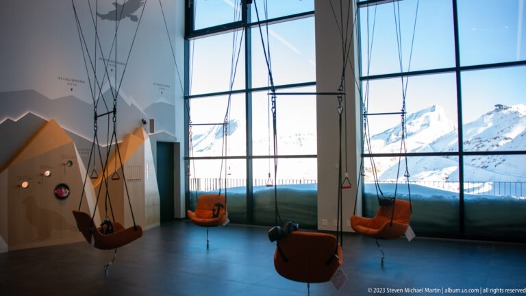 Orange suspended seats hanging inside a modern exhibit space with large windows overlooking snowy Alpine mountains.