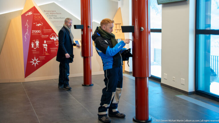 Two men using tall red interactive stations inside a modern Matterhorn-themed visitor exhibit.