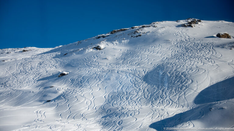 Steep snowy Alpine slope covered with ski and snowboard tracks, with exposed rock near the crest.