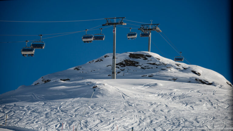 Chairlift with several suspended chairs crossing a snowy ridge beneath a deep blue sky.
