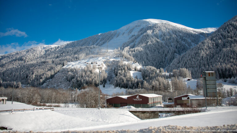 Snow-covered Alpine slope with forest, open clearings and scattered buildings beneath a deep blue sky.