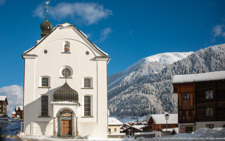 White gabled Alpine church façade with carved doorway, small canopy, round window and saint niche against snowy mountains.