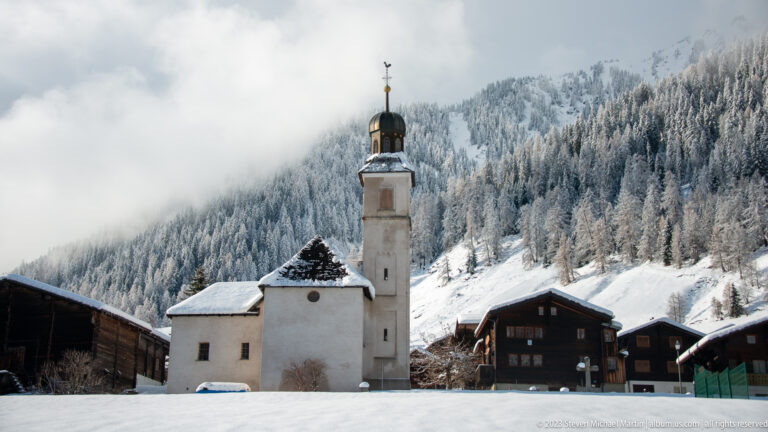 White Alpine church with tall square bell tower and dark onion-shaped roof among timber houses and snowy forest.