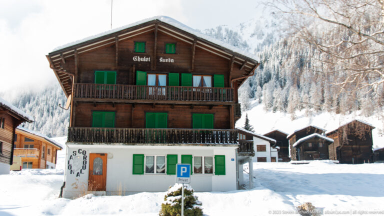 Chalet Suetta, a dark timber house with green shutters and wooden balconies above a white ground floor in deep snow.