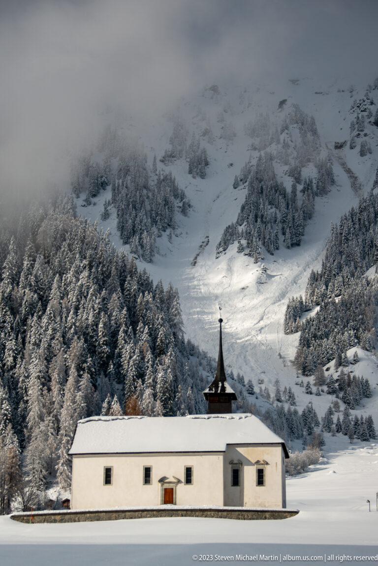 Long white chapel with a small dark roof turret standing alone in a snowfield below a steep mountain slope.