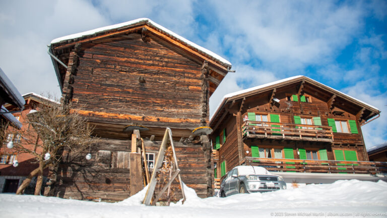 Weathered timber barn beside a larger chalet with green shutters and balconies, both surrounded by deep snow.